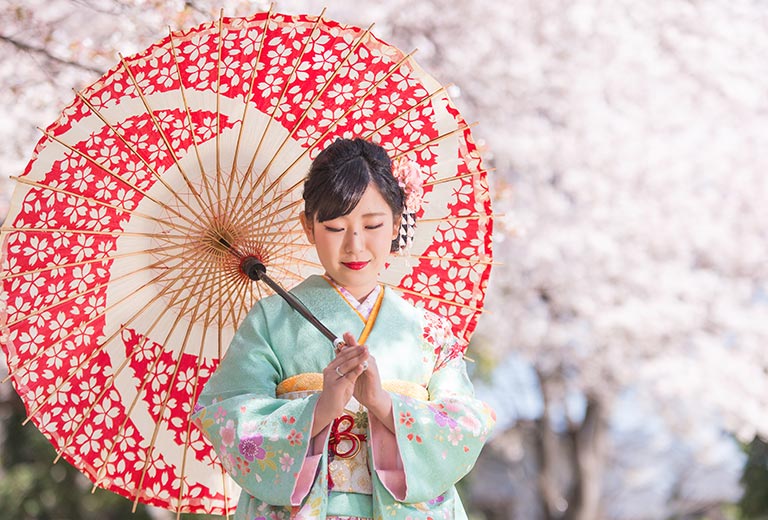 桜の木下で、振袖を着た女性が傘をさす様子